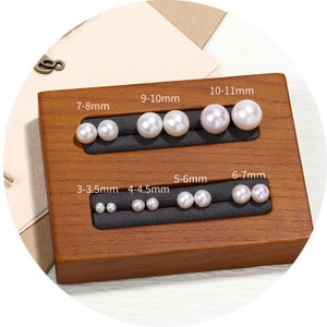 Op de afbeelding: Een houten displaybox met een verscheidenheid aan parelformaten, variërend van 3-3,5 mm tot 10-11 mm. De parels zijn wit en rond en zijn gerangschikt in rijen op de doos.