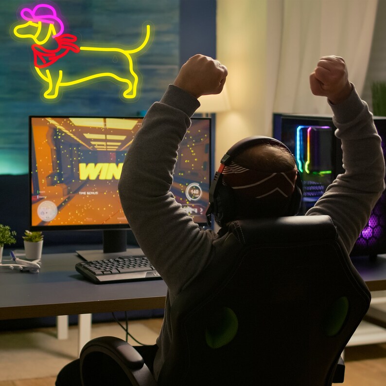 May include: A person wearing a headset sits in a gaming chair with their arms raised in victory. The person is in front of a computer monitor displaying a video game with the word "WIN" on the screen. A neon yellow dachshund with a cowboy hat and bandana is on the wall behind the person.