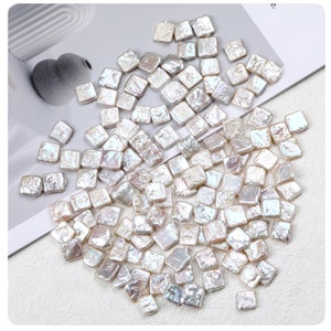 Puede incluir: Un montón grande de cuentas de perla blancas, cuadradas e irregulares. Las cuentas están esparcidas sobre una superficie blanca.