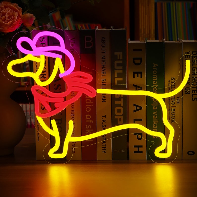 May include: A neon sign shaped like a dachshund dog wearing a pink hat. The sign is yellow with red and pink accents. The sign is lit up and is sitting on a shelf with books behind it.