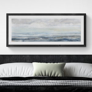 Puede incluir: Una impresión en lienzo enmarcada en negro de un paisaje marítimo con un cielo y agua azul claro, y un primer plano azul oscuro. La impresión está colgada en una pared blanca sobre una cabecera de terciopelo negro con almohadas blancas y verdes. Una pequeña planta en maceta se encuentra sobre una mesita de noche redonda blanca.