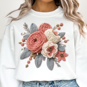 May include: White sweatshirt with a 3D floral design. The bouquet features pink, cream, and coral flowers, grey leaves, and gold accents. The sweatshirt is a comfortable, casual style.