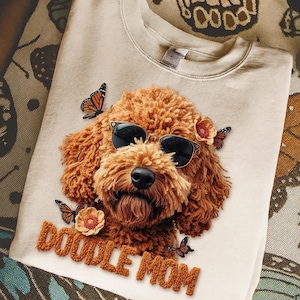 May include: Cream-coloured sweatshirt with a brown Doodle dog wearing sunglasses, butterflies, and flowers. The text "DOODLE MOM" is embroidered in brown. A comfortable and fashionable garment.