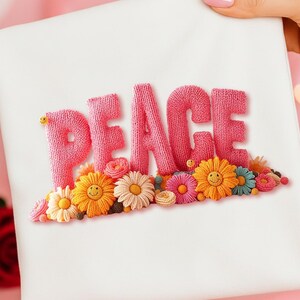 May include: A white square textile with the word "PEACE" embroidered in pink, above a row of colorful smiling flowers. The flowers include yellow, orange, pink, and blue, with green leaves. The background is a soft pink.
