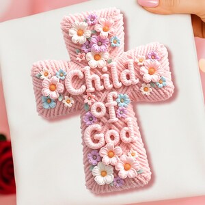 May include: A pink cross with the words "Child of God" and floral embellishments. The cross is made of a woven material and decorated with various pastel-colored flowers. The cross is on a white background.