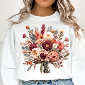 May include: White sweatshirt with a detailed floral bouquet design. The bouquet features pink, burgundy, peach, and orange flowers, with green and grey foliage. The sweatshirt is a casual, comfortable style.