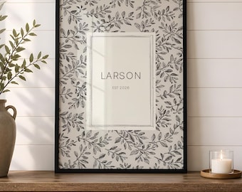 May include: A rectangular black-framed wall art piece featuring a cream-colored center panel with the text LARSON and EST 2026, surrounded by a repeating black and white botanical leaf pattern. The print is encased in a thin black frame.