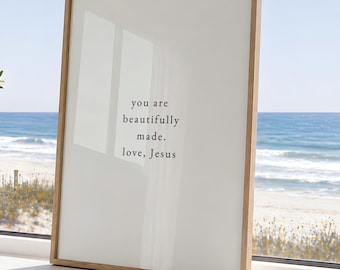 May include: A rectangular wall art print in a light wood frame featuring a white background with black text that reads: you are beautifully made. love, Jesus. The print is positioned upright on a flat surface with a clear view of the ocean.