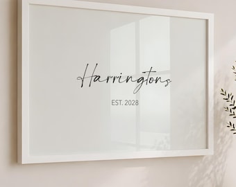 May include: A rectangular wall art piece in a white frame featuring the black text Harringtons and EST. 2028. Below the frame, a stack of books includes one with the text CALIFORNIA USA, topped by a small beige bowl, next to a tall vase with greenery.