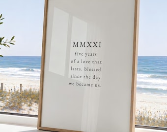 May include: A rectangular framed print with a light wood frame featuring black text on a white background that reads MMXXI five years of a love that lasts. blessed since the day we became us. The print is positioned upright on a flat surface.