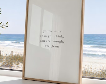 May include: A rectangular wall art print in a light wood frame featuring a white background with black text that reads: you're more than you think, you are enough. love, Jesus. The print is displayed upright on a flat surface.