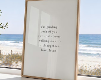 May include: A rectangular framed wall art piece featuring a light cream background with black printed text that reads: i'm guiding both of you, two soul sisters walking on this earth together. love, Jesus. The frame is made of light-colored wood.
