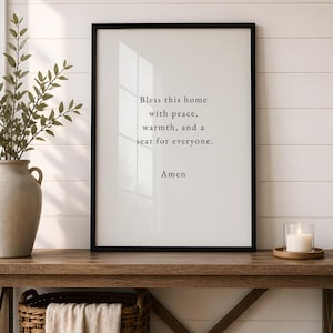 May include: A framed vertical wall art print with a white background and black text that reads: Bless this home with peace, warmth, and a seat for everyone. Amen. The print is housed in a thin black frame and displayed on a wooden console table.
