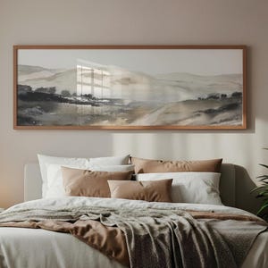 May include: A landscape art print in a wooden frame, showing rolling hills in shades of grey, beige, and brown. The artwork hangs above a bed with white bedding, brown cushions, and a textured throw. The room has a neutral colour scheme.