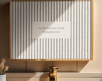 May include: A rectangular wall art piece in a gold frame featuring a white background with vertical grey stripes and the text "CUTE HANDS WASH THEMSELVES" centered in a white box. The artwork is positioned above a gold faucet and sink.