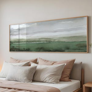 Peut inclure: Une peinture de paysage à l'aquarelle encadrée, dans des tons de vert et de gris, est accrochée au-dessus d'un lit avec des oreillers et une couverture. L'œuvre d'art est rectangulaire avec un cadre en bois clair, et le lit a une palette de couleurs neutres.