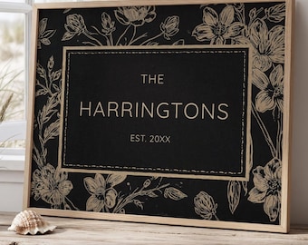 May include: A framed family name sign with a black background and floral design. The sign reads "THE HARRINGTONS" with "EST. 20XX" below. The frame is a light wood color, and the floral design is a light tan color.