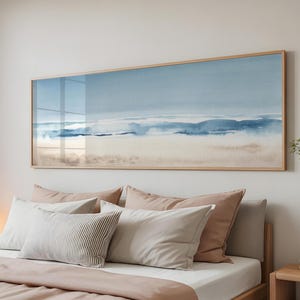 May include: A large rectangular framed watercolor painting depicting a coastal landscape in shades of blue, beige, and white. The artwork hangs above a bed with various pillows and a blush-colored duvet cover. The frame is made of light-colored wood.