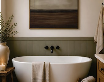 May include: A white freestanding oval bathtub centered against a sage green paneled wall. Above the tub hangs a landscape painting in a gold frame. A beige towel with fringe hangs over the tub edge, and a wicker basket holds a rolled towel nearby.