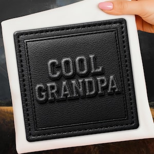 May include: Black leather patch with the words "COOL GRANDPA" embossed in a bold, textured font. The patch is rectangular with a stitched border and is set against a white background.