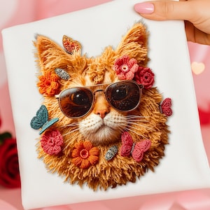 Op de afbeelding: Een wit vierkant kussen met een gedetailleerd oranje kattengezicht met een zonnebril. De kat is versierd met gehaakte bloemen en vlinders in rood, oranje en blauwgroen. Het kussen wordt vastgehouden door een persoon met roze nagellak.