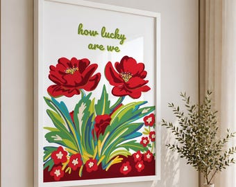 May include: A framed wall art print featuring two large red flowers with yellow centers and green foliage. Below the flowers are smaller red blossoms with white centers. The text "how lucky are we" is printed in green at the top of the white background.