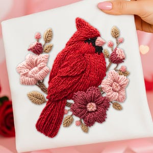 May include: Embroidered square featuring a vibrant red cardinal bird surrounded by flowers in shades of pink and burgundy, with tan leaves. The bird has a black face and a pink beak. The artwork is on a white background.