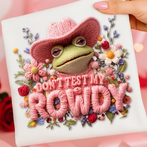 Könnte beinhalten: Ein weißes Quadrat mit einem detaillierten Stickdesign, das einen Frosch mit einem rosa Cowboyhut und dem Text "DON'T TEST MY ROWDY" zeigt. Der Frosch ist von gestickten Blumen in verschiedenen Farben umgeben.