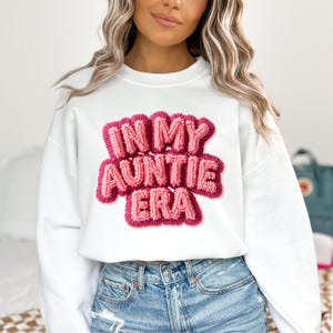 May include: White sweatshirt with the phrase "IN MY AUNTIE ERA" in a pink, textured design. The sweatshirt is a crew neck style and is paired with blue denim jeans.