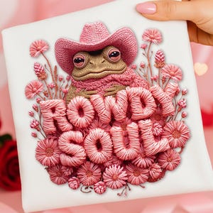 Op de afbeelding: Een vierkant wit canvas toont een grillig ontwerp van een kikker met een roze cowboyhoed en trui. De tekst "ROWDY SOUL" is geborduurd met roze draad, omringd door roze bloemen.