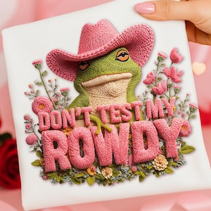 May include: White square with a detailed embroidered design. A green frog wearing a pink cowboy hat is centered, with the text "DON'T TEST MY ROWDY" in pink, surrounded by pink flowers and greenery.