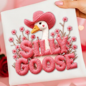 Könnte beinhalten: Ein weißes Quadrat mit einem skurrilen Stickdesign. Es zeigt eine Gans mit einem rosa Cowboyhut, umgeben von rosa Blumen und den Worten "Silly Goose" in rosa Faden. Die Gans hat einen gelben Schnabel und ein Herz auf der Brust.