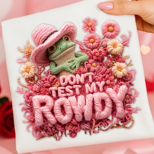 Könnte beinhalten: Ein weißes Quadrat mit einem detaillierten Stickdesign. Ein grüner Frosch mit einem rosa Cowboyhut ist von rosa Blumen umgeben. Der Text "DON'T TEST MY ROWDY" ist mit rosa Garn gestickt.