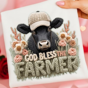 May include: A white square decorative pillow featuring a black cow wearing a beige cap, surrounded by embroidered flowers and the text "GOD BLESS THE FARMER" in a textured font. The design has a 3D effect.