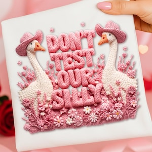 Könnte beinhalten: Ein weißes quadratisches Kissen mit einem Strickdesign mit zwei Gänsen mit rosa Hüten, umgeben von rosa Blumen. Der Text "DON'T TEST OUR SILLY" ist in rosa Strickbuchstaben.