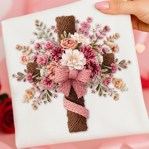 May include: A white square canvas featuring a brown rope cross adorned with a floral arrangement. The flowers are in shades of pink, peach, and white, with a pink knitted bow at the center. Green leaves and small flowers complete the design.