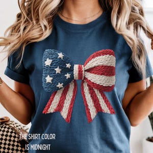 以下が含まれることがあります： 紺色のTシャツ。赤、白、青の大きなリボン模様で、青い部分には白い星が描かれています。リボンはシャツの中央に配置されています。シャツの下部には「THE SHIRT COLOR IS MIDNIGHT」と書かれています。