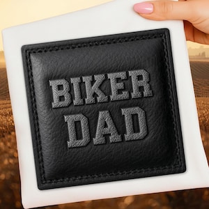 May include: A black leather patch with the words "BIKER DAD" embossed in a bold, gray font. The patch is square with a stitched border and is set against a white background.