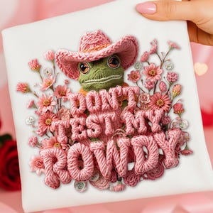 Puede incluir: Un lienzo cuadrado blanco con un diseño caprichoso. Presenta una rana verde con un sombrero de vaquero rosa, rodeada de flores rosas. El texto "DON'T TEST MY ROWDY" se muestra prominentemente en hilo rosa.