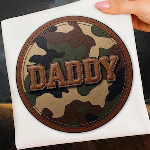 May include: A white square item with a circular patch featuring a camouflage pattern in shades of brown, green, and tan. The word "DADDY" is displayed in brown, block letters with a leather-like texture.