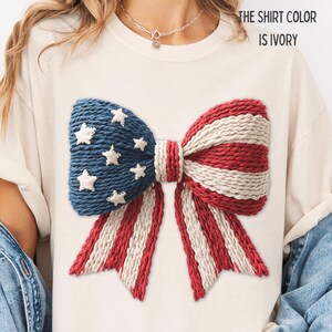 以下が含まれることがあります： 大きなリボンデザインのアイボリー色のTシャツ。リボンは青、赤、白のテクスチャード加工された生地で作られており、青い部分には白い星が付いています。「THE SHIRT COLOR IS IVORY」というテキストが上部に印刷されています。