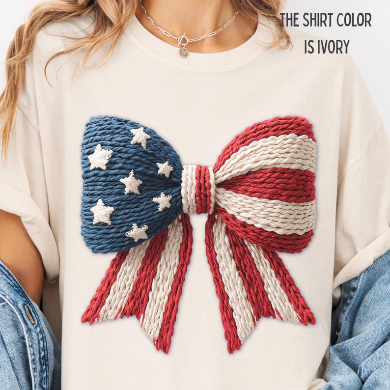 以下が含まれることがあります： 大きなリボンデザインのアイボリー色のTシャツ。リボンは青、赤、白のテクスチャード加工された生地で作られており、青い部分には白い星が付いています。「THE SHIRT COLOR IS IVORY」というテキストが上部に印刷されています。