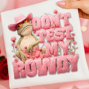 Könnte beinhalten: Weißes T-Shirt mit einer gestrickten Froschfigur, die einen rosa Cowboyhut und ein Kleid trägt. Der Schriftzug "DON'T TEST MY ROWDY" ist in rosa Garn gestickt, mit floralen Akzenten. Der Frosch hat orangefarbene Augen und hält ein rosa Accessoire.
