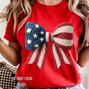 以下が含まれることがあります： 愛国的なリボンデザインの赤いTシャツ。リボンは、白い星が付いた青い部分と、赤と白のストライプの部分があります。シャツの下部にあるテキストは「シャツの色は赤です」と書かれています。