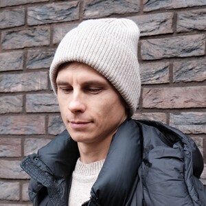 K&ouml;nnte beinhalten: Ein Mann tr&auml;gt eine beige Strickm&uuml;tze und eine schwarze Daunenjacke mit Kapuze. Die Jacke hat einen Rei&szlig;verschluss und Kn&ouml;pfe.