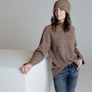 May include: A woman wearing a brown turtleneck sweater and blue jeans. She is wearing a brown knit beanie and is leaning against a white cube.