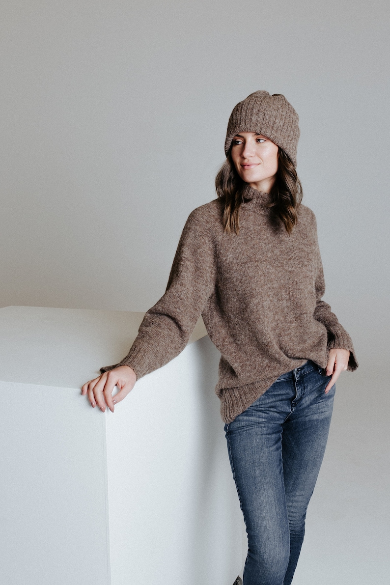 May include: A woman wearing a brown turtleneck sweater and blue jeans. She is wearing a brown knit beanie and is leaning against a white cube.