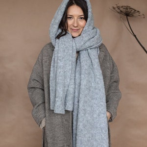 May include: A woman wearing a gray coat and a light blue fuzzy scarf. The scarf is wrapped around her neck and head, and she is looking at the camera.