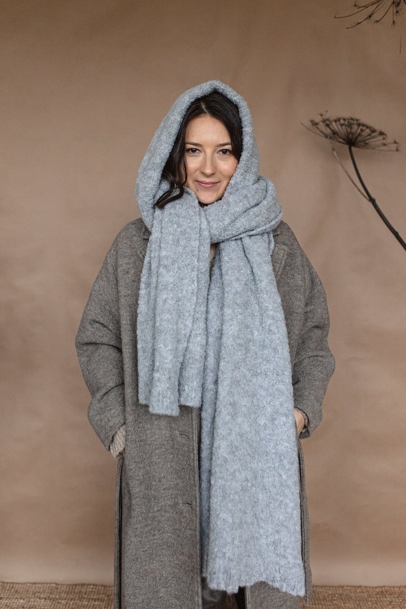 May include: A woman wearing a gray coat and a light blue fuzzy scarf. The scarf is wrapped around her neck and head, and she is looking at the camera.