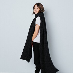 May include: A woman wearing a black fuzzy scarf, a white t-shirt, black pants, and black boots. She is standing in front of a light blue wall.
